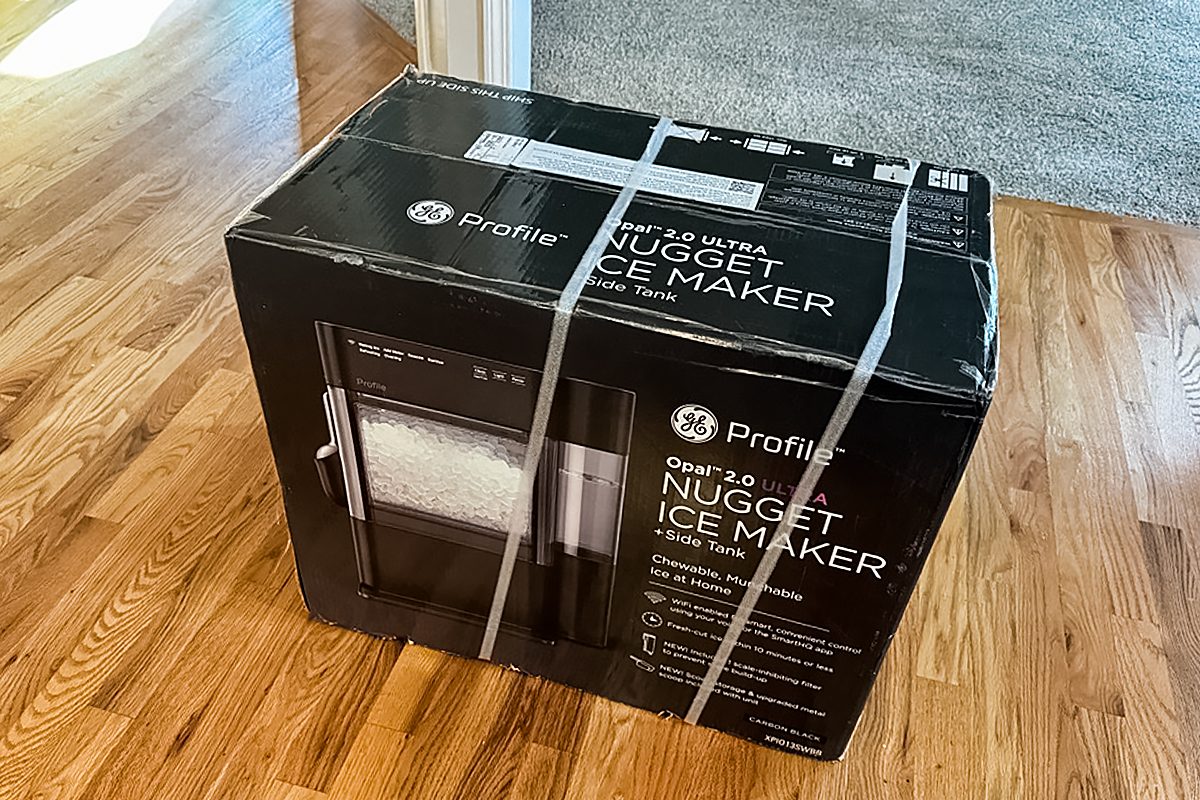 A sealed black box for a GE Profile Opal 2.0 Nugget Ice Maker sits on a wooden floor, secured with white plastic straps.