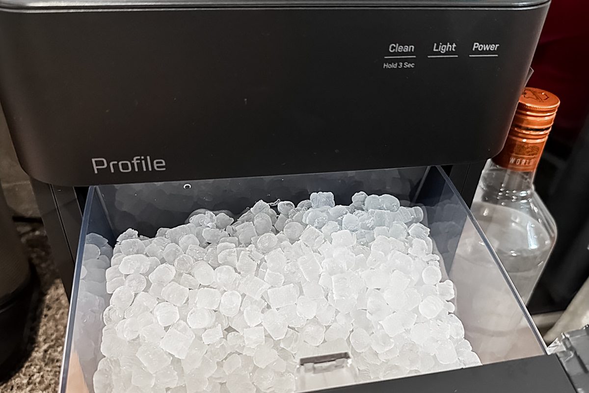 A countertop ice maker filled with small, clear ice nuggets. The control panel displays buttons labeled Clean, Light, and Power. A bottle with a copper-colored cap stands next to the machine.