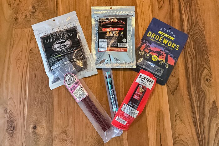Five different packaged meat snacks, including beef jerky and sausage sticks, are arranged in a fan shape on a wooden surface. The packages feature various labels and colors.