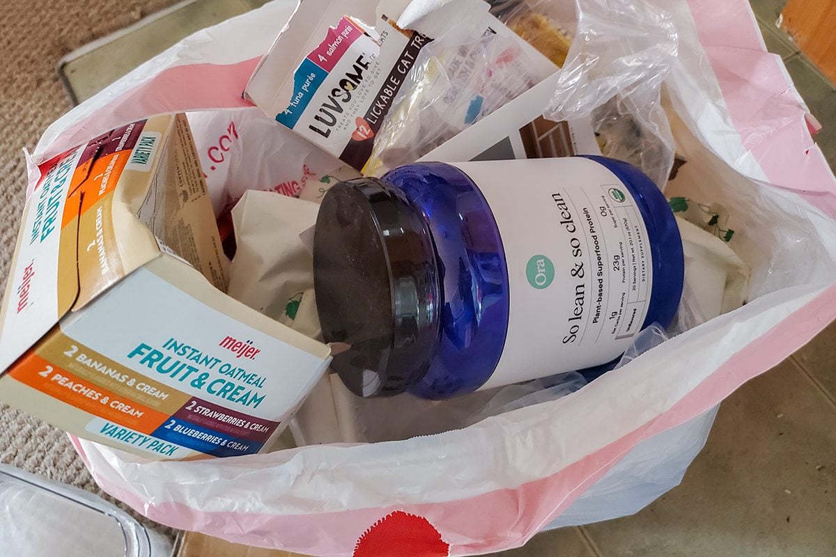 A close-up of a shopping bag containing a blue-labeled vitamin jar, a box of fruit and grain breakfast bars, and various other food and health items in their packaging.