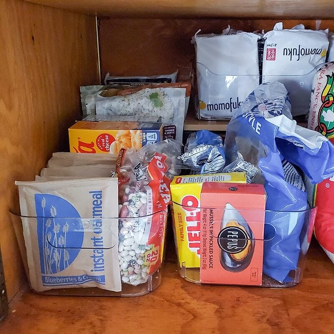 A kitchen cabinet with organized dry goods, including rice, cereal, tea, oatmeal packets, and other packaged foods, neatly stored in clear bins on a wooden shelf.