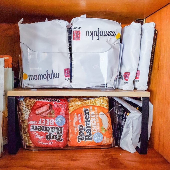 A wooden pantry shelf holds packets of Momofuku noodles on the top and various instant noodles, including Top Ramen and Tapatío Hot, stacked below.