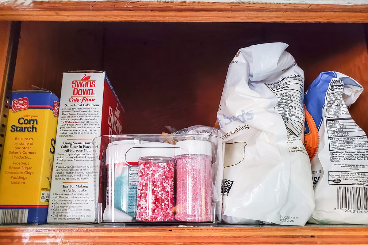 A kitchen cabinet shelf with boxes of cornstarch and cake flour, a bag of sugar, a bag of flour, and a clear container holding small jars of pink and white sprinkles and other baking decorations.