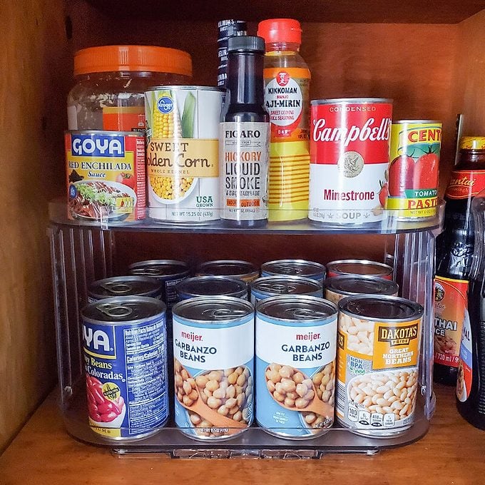 A pantry shelf with canned goods, including beans, corn, Campbell’s minestrone soup, tomato paste, and condiments such as soy sauce and lemon juice, neatly organized on two clear tiers.