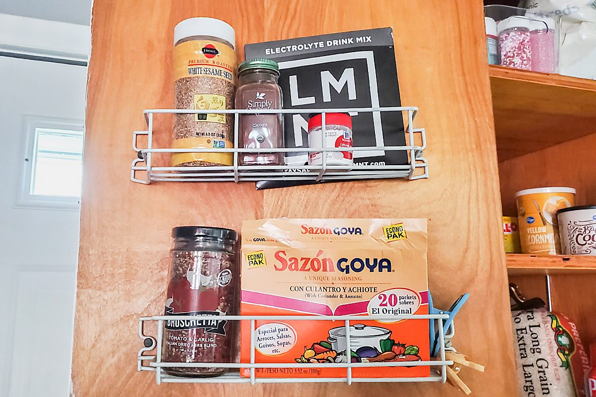 Two wire racks attached to a wooden cabinet door hold various items, including seasonings, a jar, an LM electrolyte drink mix packet, and a box of Sazón Goya seasoning.