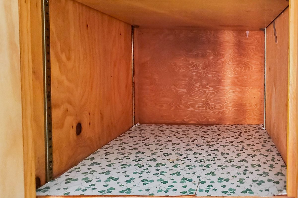The image shows the inside of an empty wooden cabinet. The bottom surface is lined with white paper decorated with a pattern of green clovers. The cabinet walls and ceiling are plain wood.