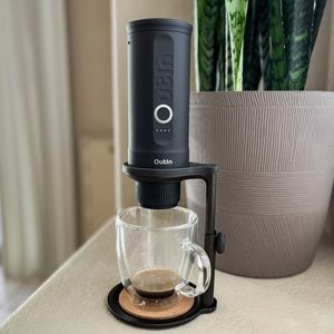 A compact black coffee maker sits above a clear glass mug on a stand, brewing coffee, next to a large textured planter with tall green plants. The setup is placed on a light-colored surface indoors.