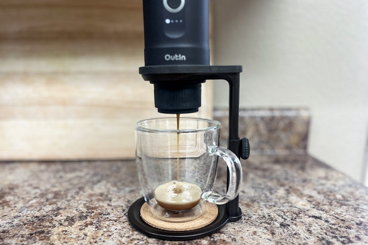 A countertop espresso machine dispenses coffee into a clear glass mug, producing a layer of crema on top. The machine is black and labeled "Outin." The setup is on a speckled kitchen counter with a light backdrop.