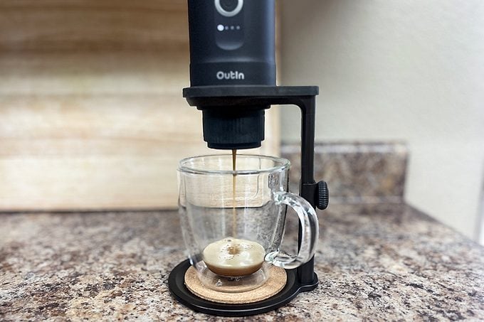A countertop espresso machine dispenses coffee into a clear glass mug, producing a layer of crema on top. The machine is black and labeled "Outin." The setup is on a speckled kitchen counter with a light backdrop.