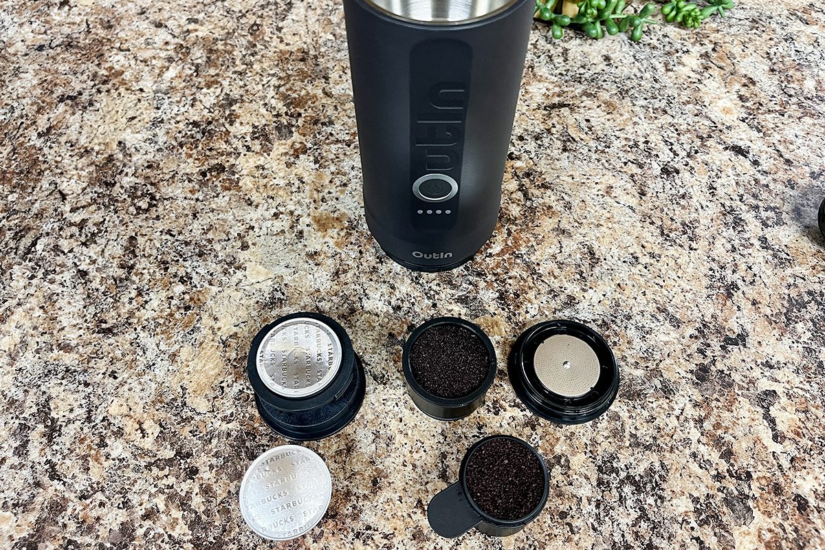 A portable coffee maker is on a speckled countertop, surrounded by various removable parts and coffee grounds, including filters and scoops, arranged neatly in front of the machine.
