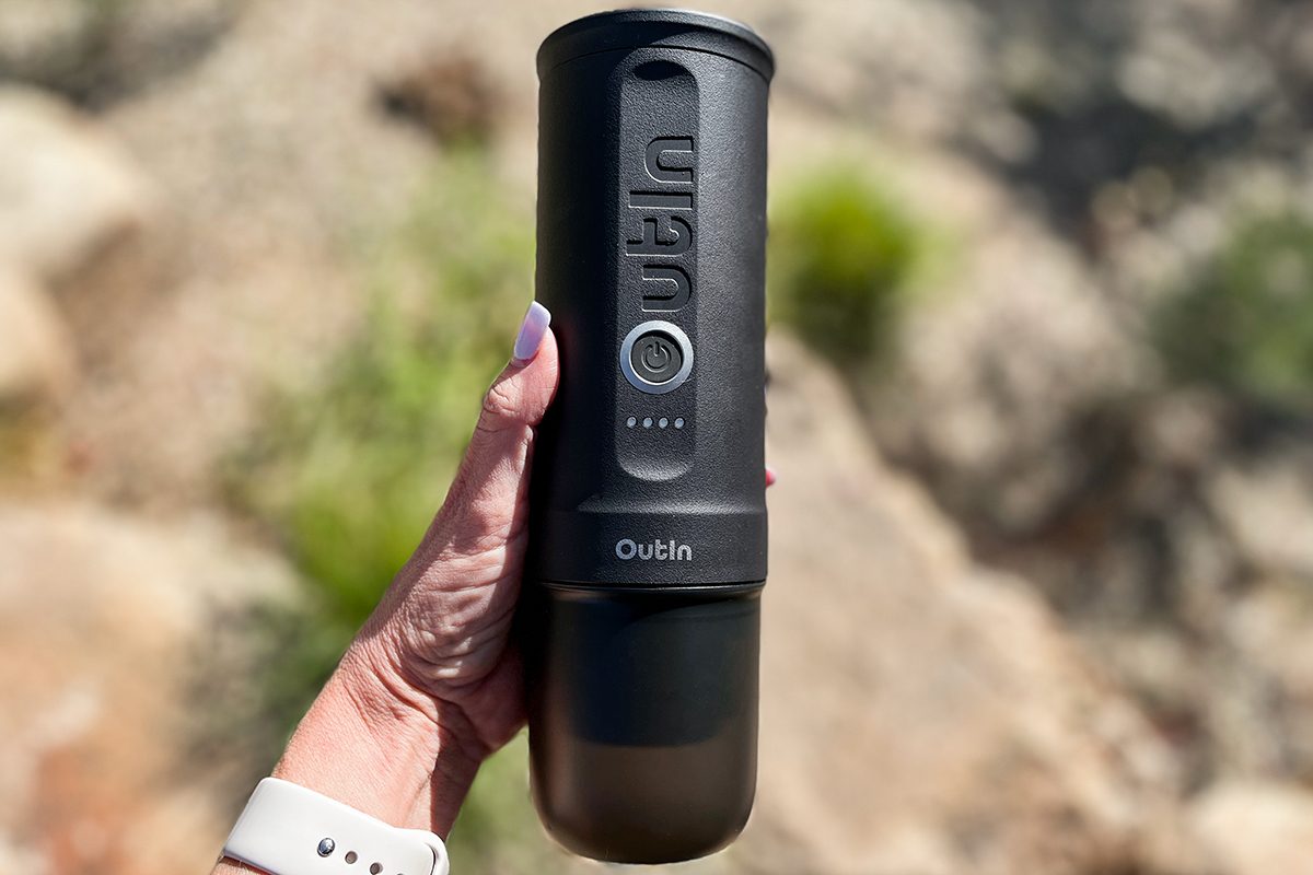 A hand holding a black Outin branded portable coffee maker outdoors, with blurred rocks and greenery in the background.