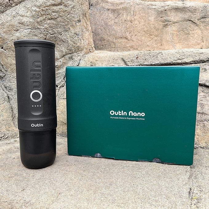 A black Outin Nano portable espresso machine stands next to a teal Outin Nano box, both placed on stone steps.