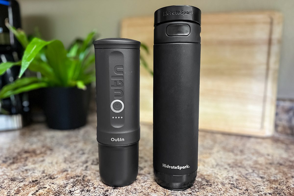 Two black travel mugs sit on a kitchen counter. The shorter one on the left has buttons and the brand name "Owala," while the taller one on the right is labeled "Hydro Flask." A plant and cutting board are in the background.