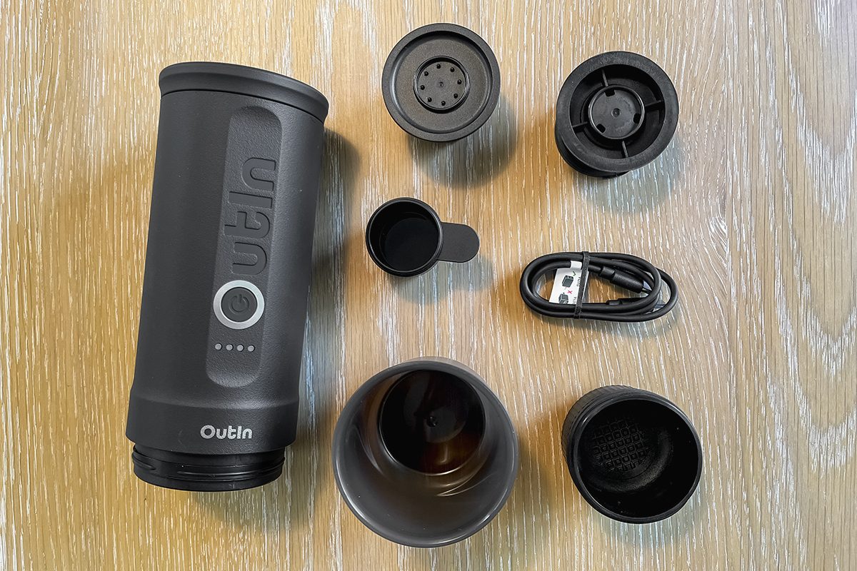 A portable electric coffee maker with detachable parts, a USB charging cable, and several lids and components arranged on a wooden surface.