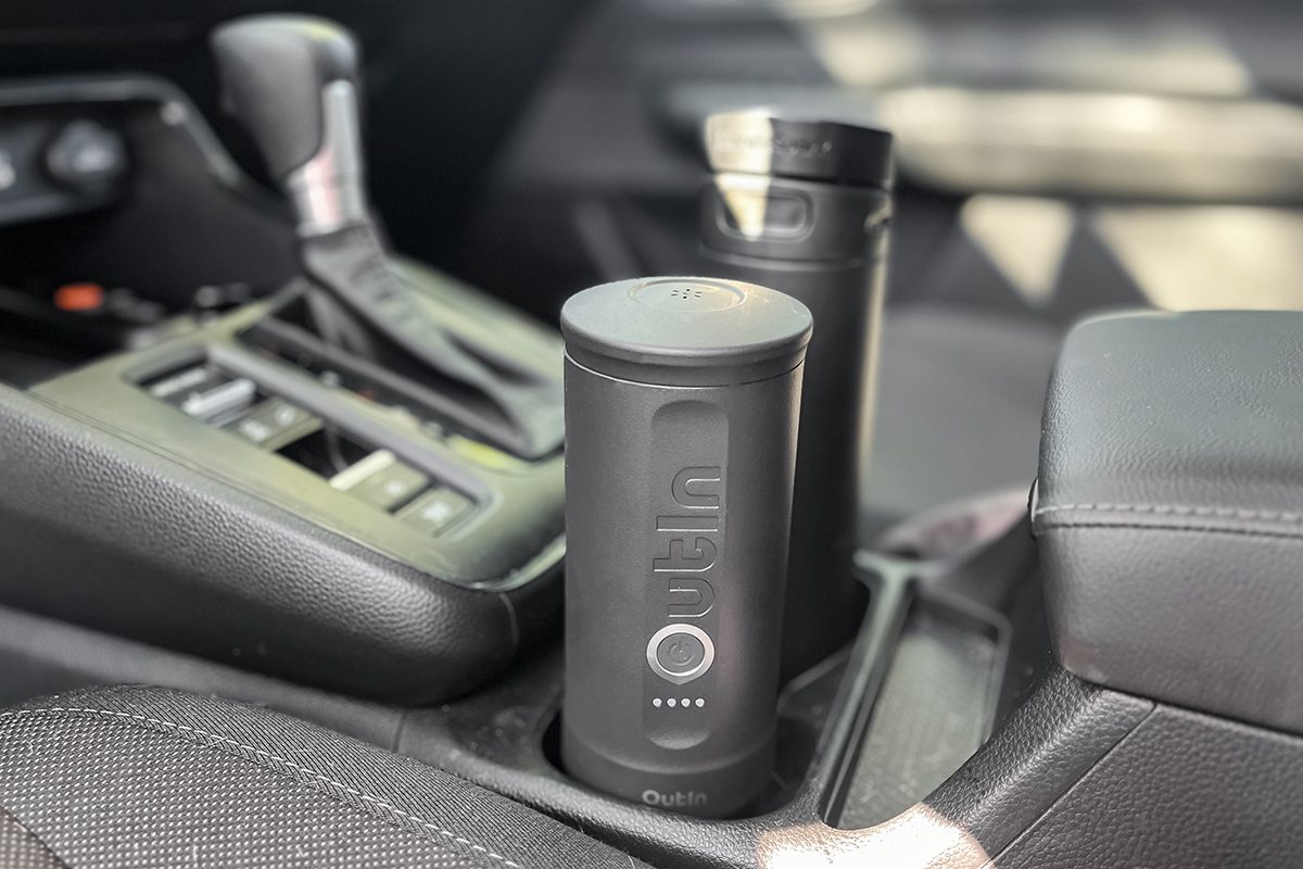 A black cylindrical travel mug labeled "Ottin" sits in the cup holder of a car next to another black travel mug, both in the vehicle's center console.