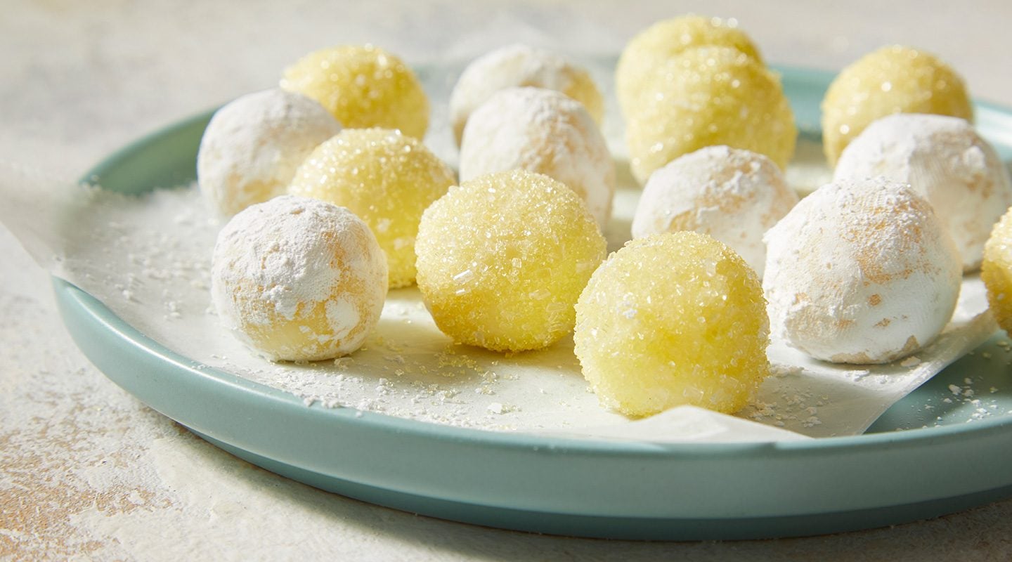 A plate of round, yellow and white sugar-coated dessert balls is displayed on a light blue dish lined with parchment paper. Some balls are dusted with powdered sugar, while others have a sparkly sugar coating.