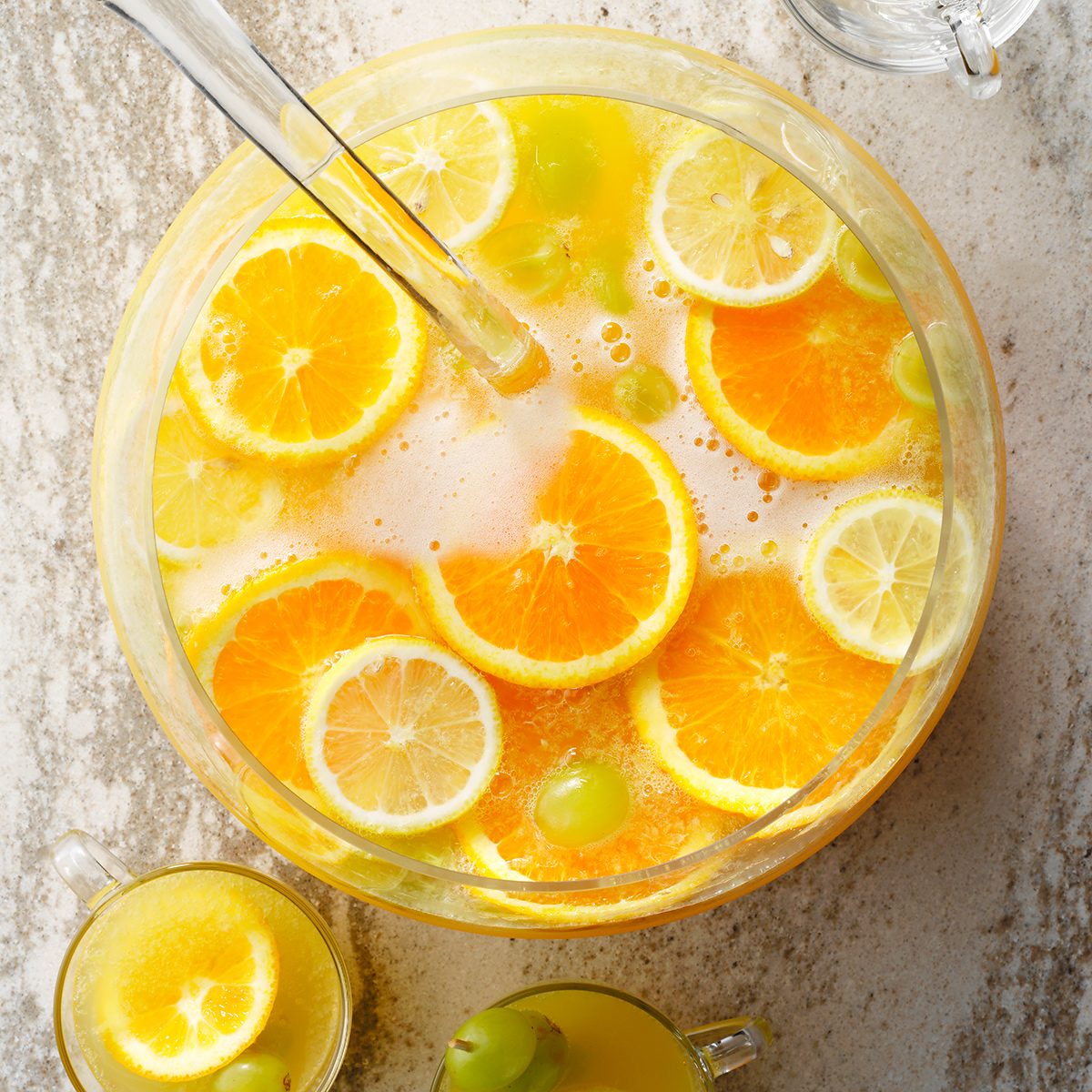 A glass punch bowl filled with a citrus drink, garnished with orange and lemon slices and green grapes, sits on a light-colored surface with a ladle inside. A small glass of the punch is beside the bowl.