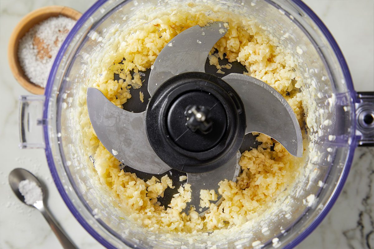 Overhead horizontal image of minced garlic in a food processor with the blade visible. To the left are small bowl and spoon holding salt.
