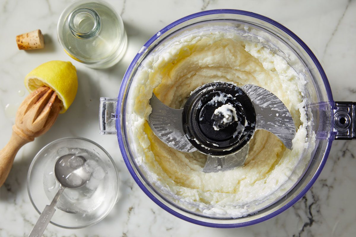 Overhead horizontal image of a food processor containing a light colored mixture, possibly aioli or mayonnaise. The mixture coats the blades of the food processor. A halved lemon in a wooden citrus juicer, a small bottle of clear liquid with a cork stopper, and a glass containing ice and a measuring spoon are nearby.