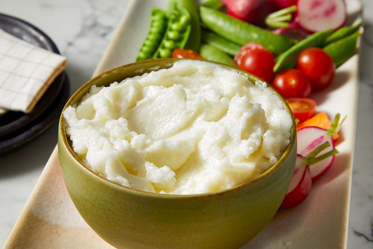 Eye Level horizontal image of a bowl filled with a white dip, sitting on a platter next to raw vegetables. The platter has tomatoes, sugar snap peas, radishes, and carrots.