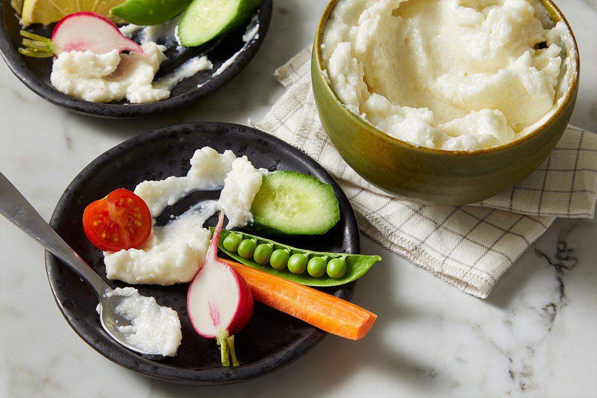 Overhead horizontal image of a bowl of white dip next to two black plates of assorted vegetables. One plate features a sliced tomato, radish, sliced cucumber, and a spoonful of the white dip, while the other plate features a lemon wedge, lime wedge, radish, sliced cucumber and dip.