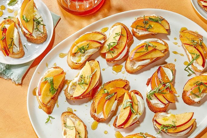 Horizontal overhead image featuring a large serving platter filled with peach crostinis topped with whipped cottage cheese and hot honey. Fresh basil and lemon zest add brightness, while a warm-toned background enhances the premium set styling and entertaining appeal.