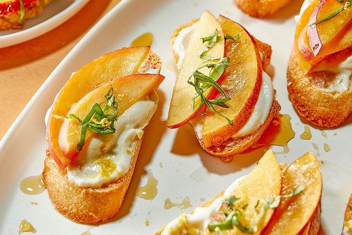 Horizontal close-up image highlighting individual crostinis topped with whipped cottage cheese, sliced peaches, basil and hot honey drizzle. The tight composition emphasizes texture—from the crisp baguette base to the creamy cheese and juicy peaches.