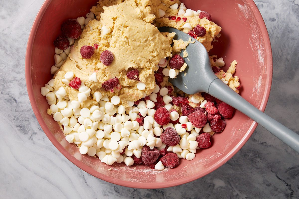 Overhead horizontal image of raspberries and white chocolate chips being folded into cookie dough.
