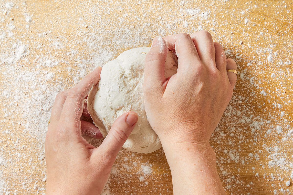 Overhead horizontal image of a person kneading dough on a wooden surface dusted with flour. The dough is centered and hands are pushing into the dough.