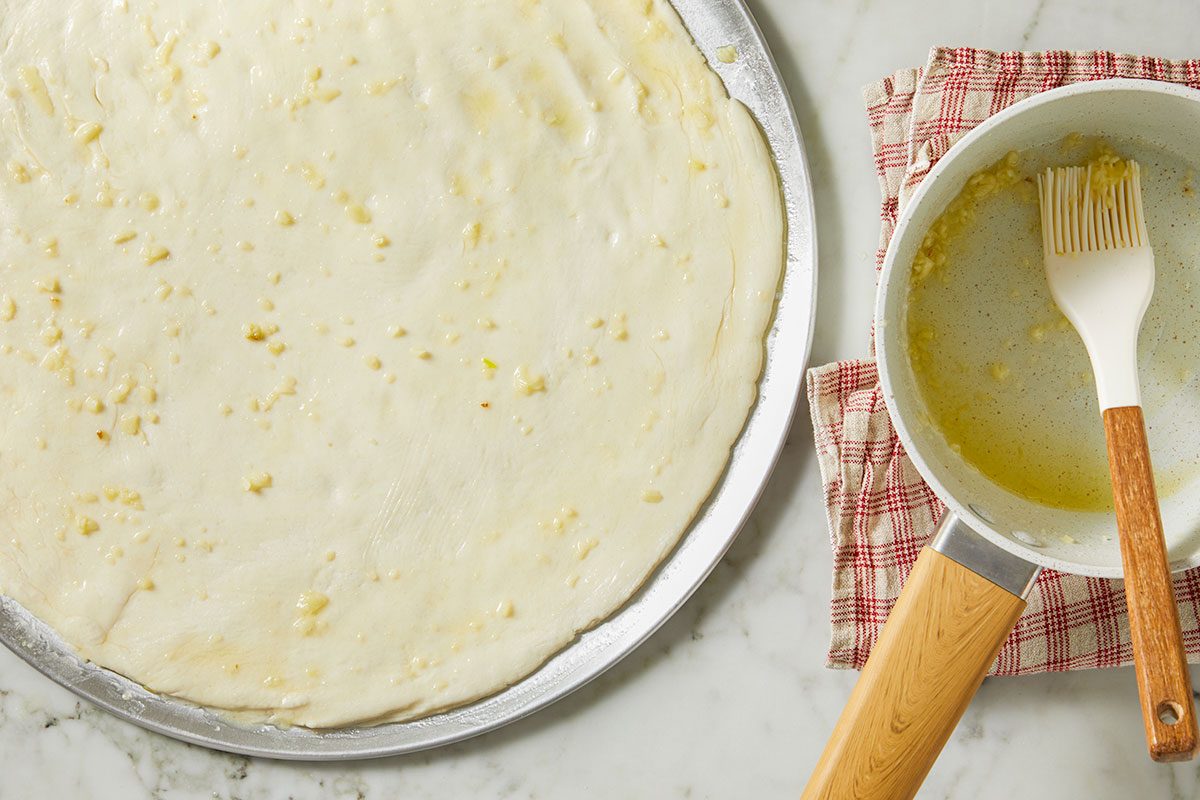 Overhead horizontal image of pizza dough spread out on a pizza pan, topped with butter and garlic. Next to the pan, there is a pot with melted butter and minced garlic in it along with a white basting brush. | [Original] Overhead horizontal image of pizza dough on a metal baking sheet, brushed with melted butter and garlic. Next to the dough is a small saucepan with a pastry brush, holding the remaining butter and garlic mixture.
