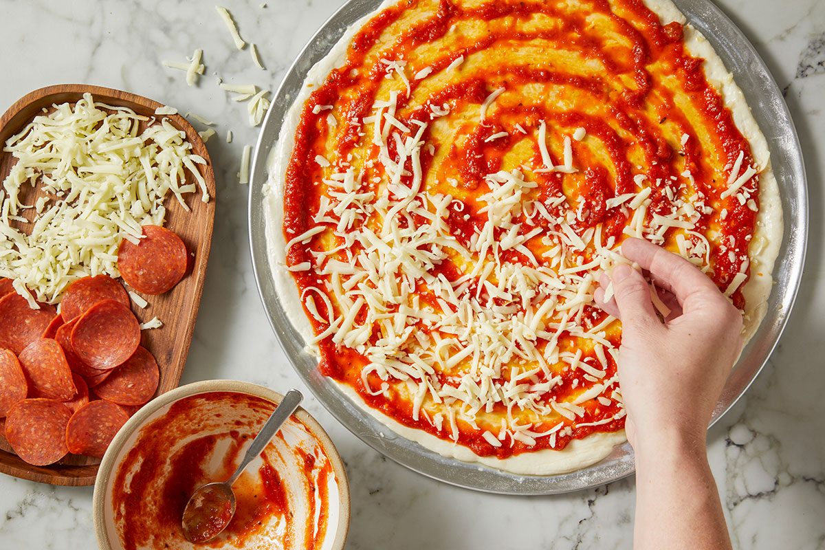 Overhead horizontal image of a person adding shredded mozzarella cheese to a pizza. The pizza dough is spread with tomato sauce, and pepperoni slices are visible in a wooden tray to the side.