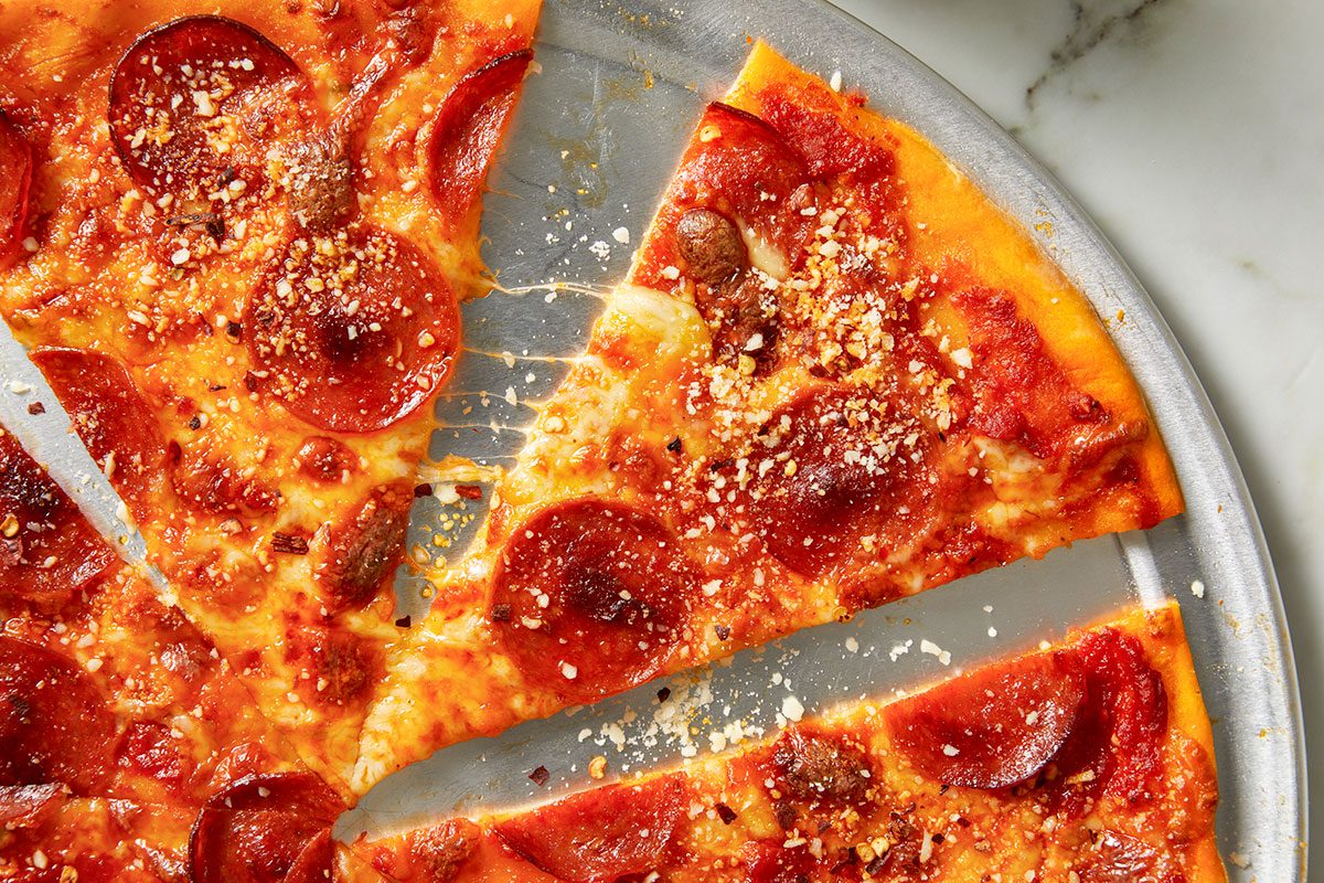 Overhead horizontal image of pepperoni pizza slices arranged on a metal tray. The pizza is topped with cheese, pepperoni, and sprinkled with parmesan cheese and red pepper flakes, some of which are visible in nearby containers.
