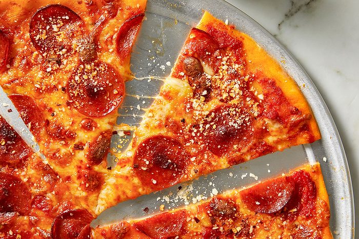 Overhead horizontal image of pepperoni pizza slices arranged on a metal tray. The pizza is topped with cheese, pepperoni, and sprinkled with parmesan cheese and red pepper flakes, some of which are visible in nearby containers.