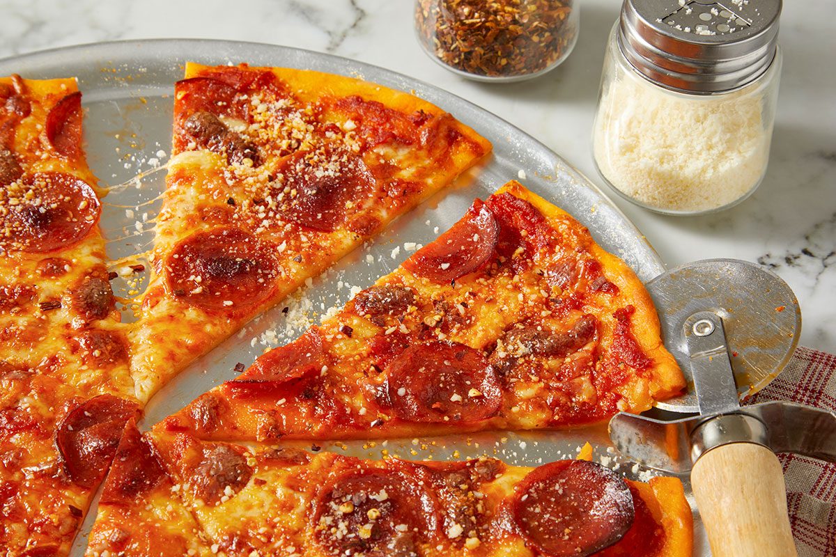 Overhead horizontal image of pepperoni pizza slices arranged on a metal tray. The pizza is topped with cheese, pepperoni, and sprinkled with parmesan cheese and red pepper flakes, some of which are visible in nearby containers.