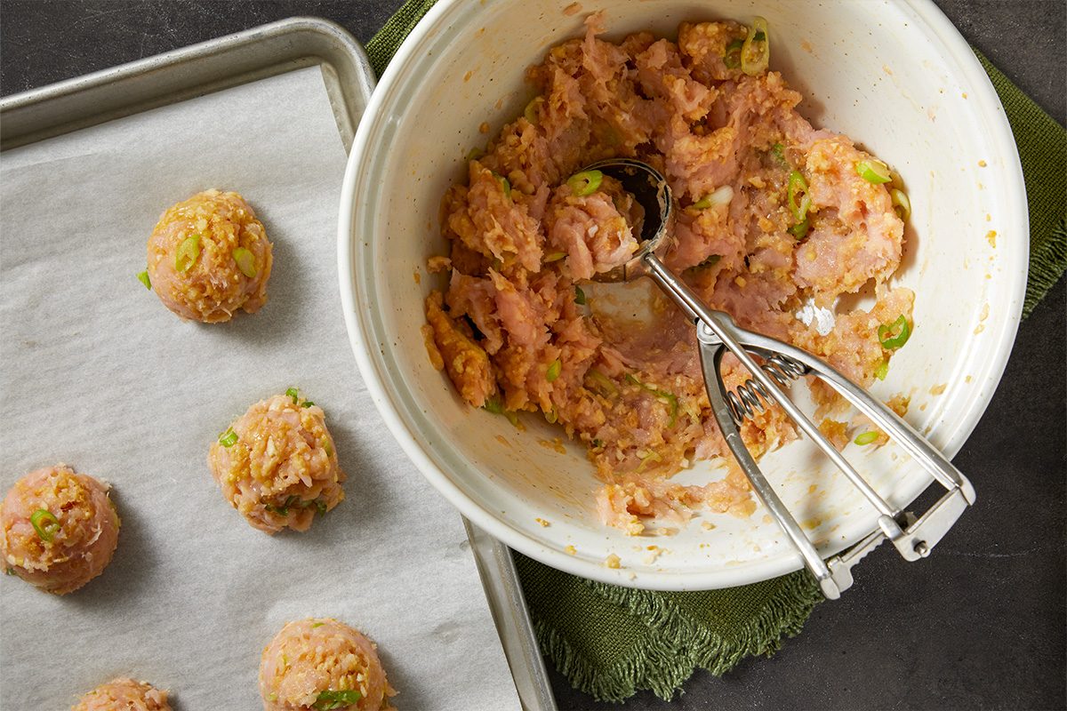 Overhead horizontal image of the next step of meatball prep. A metal scoop is in a mixing bowl with mixed ingredients. Formed meatballs are on a sheet pan lined with parchment paper.