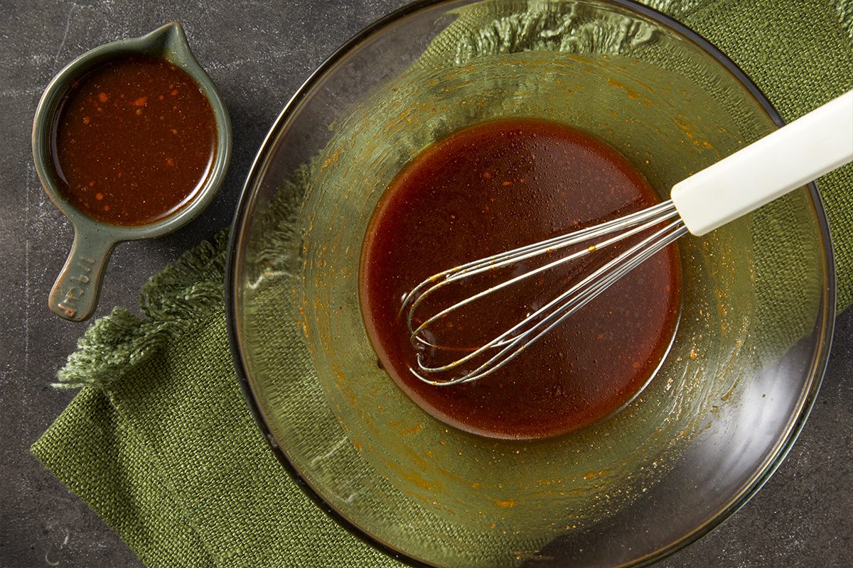 Overhead horizontal image of a clear glass bowl filled with a dark brown sauce or marinade, with a whisk resting in the mixture. A small serving pitcher containing the same liquid sits to the side, set against a green woven mat.