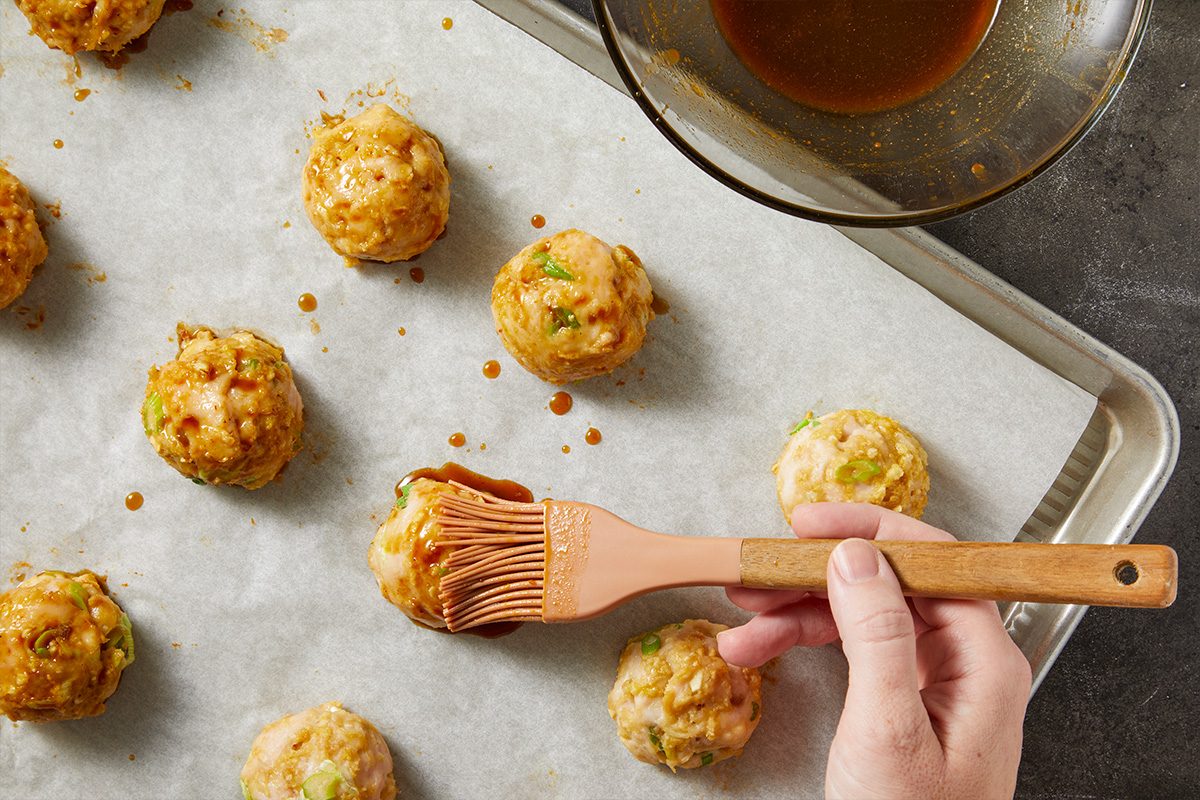 Overhead horizontal image of meatballs on a baking sheet. A hand is brushing sauce onto a meatball on the baking sheet next to a bowl of sauce.