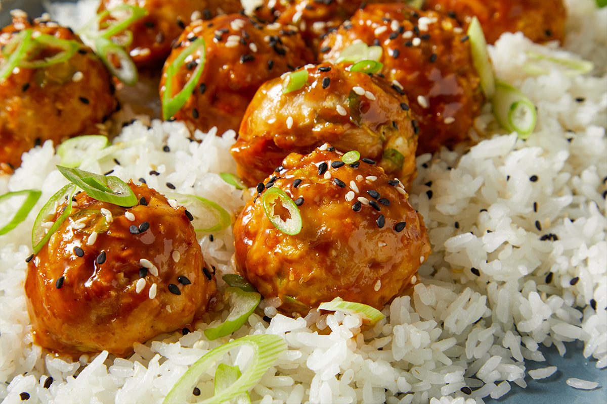 Eye Level horizontal image of a plate of glazed meatballs served over rice and garnished with sliced scallions and sesame seeds. The dish is plated and ready to eat.