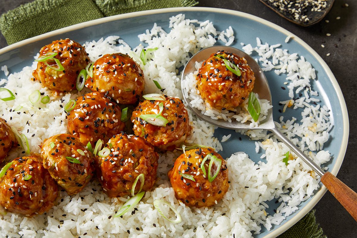 Overhead horizontal image of glazed meatballs sprinkled with black and white sesame seeds, on a bed of fluffy white rice and garnished with sliced green onions. A serving spoon holds one meatball.