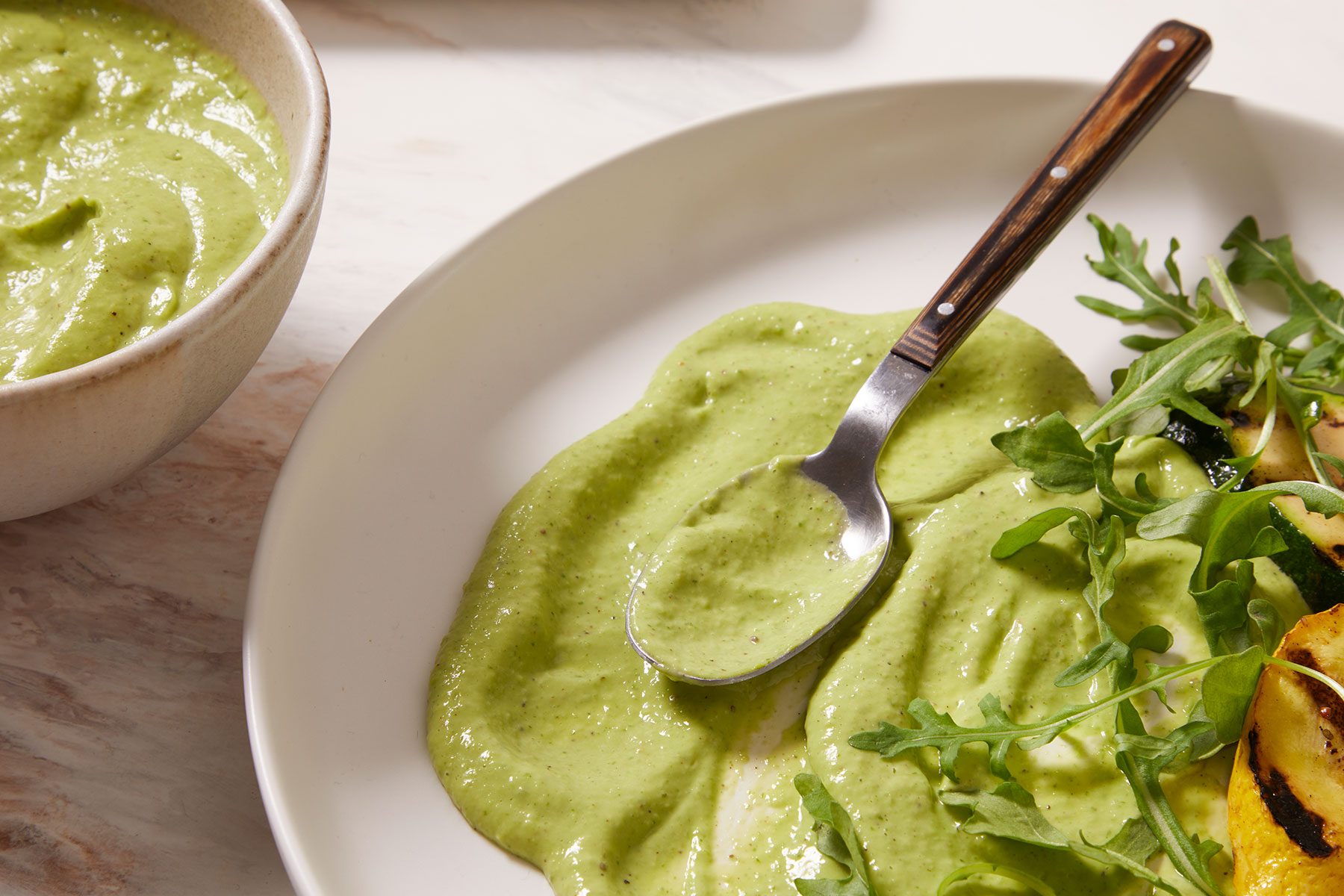Avocado Basil Dressing spread on a white plate with some more in a bowl placed on side. A spoon in the dip with some greens on it.