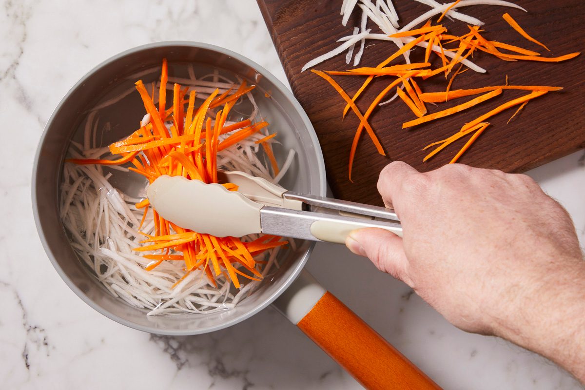 Overhead horizontal image of julienned carrots and daikon radish being transferred with tongs from a pot of water to a wooden cutting board. The hand of the person using the tongs is visible in the frame.