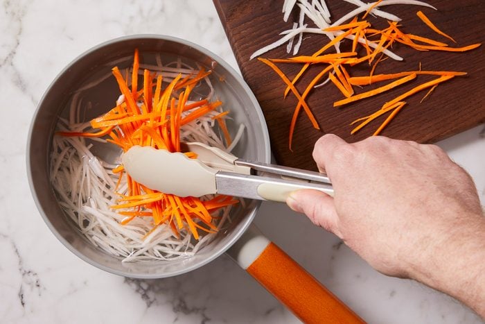 Overhead horizontal image of julienned carrots and daikon radish being transferred with tongs from a pot of water to a wooden cutting board. The hand of the person using the tongs is visible in the frame.