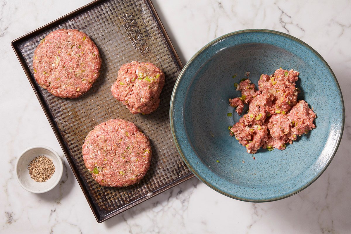 Overhead horizontal image of a blue ceramic bowl filled with uncooked ground meat and green onions next to a metal baking sheet that has two patties already formed and a third portion of the mixture. A bowl with pepper is also visible.