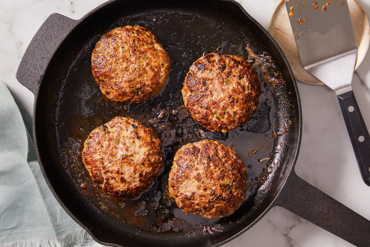 Overhead horizontal image of four burger patties cooking in a cast iron skillet. A metal spatula sits on the right side of the pan, on top of a wooden board.