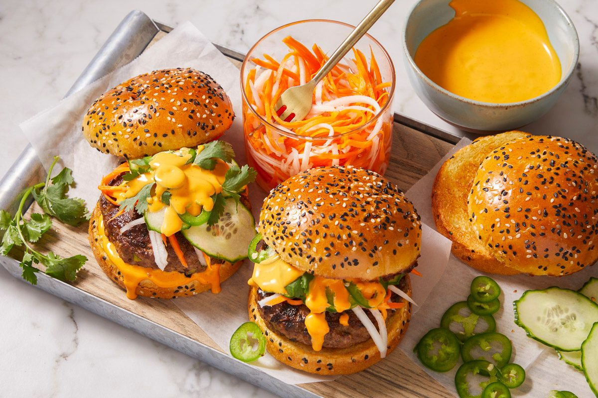 Overhead horizontal image of burgers on a metal tray topped with sauce, cilantro, carrots, cucumbers and jalapenos. A container of carrots and radishes and a bowl of sauce also sit on the tray.