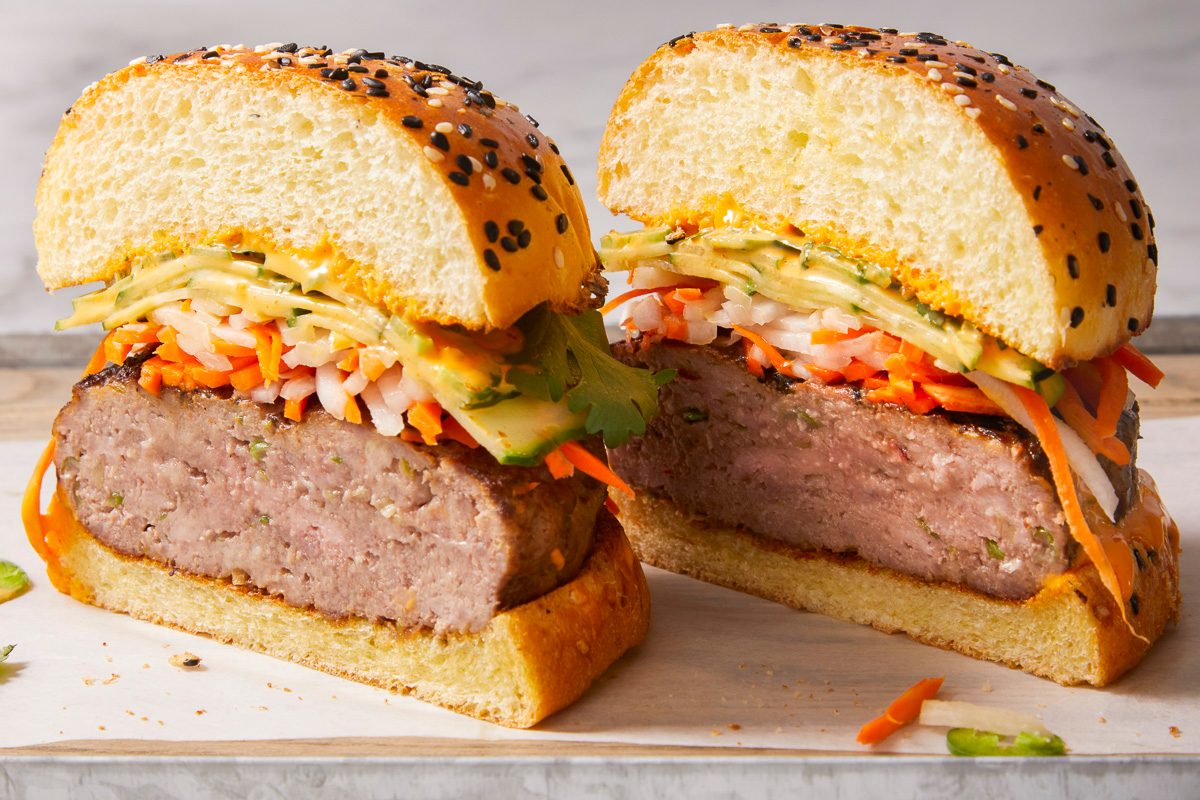 Eye Level horizontal image of a cut burger. The halves of the burger show shredded carrots, onions, zucchini, and a sauce between the patty and the seeded bun.