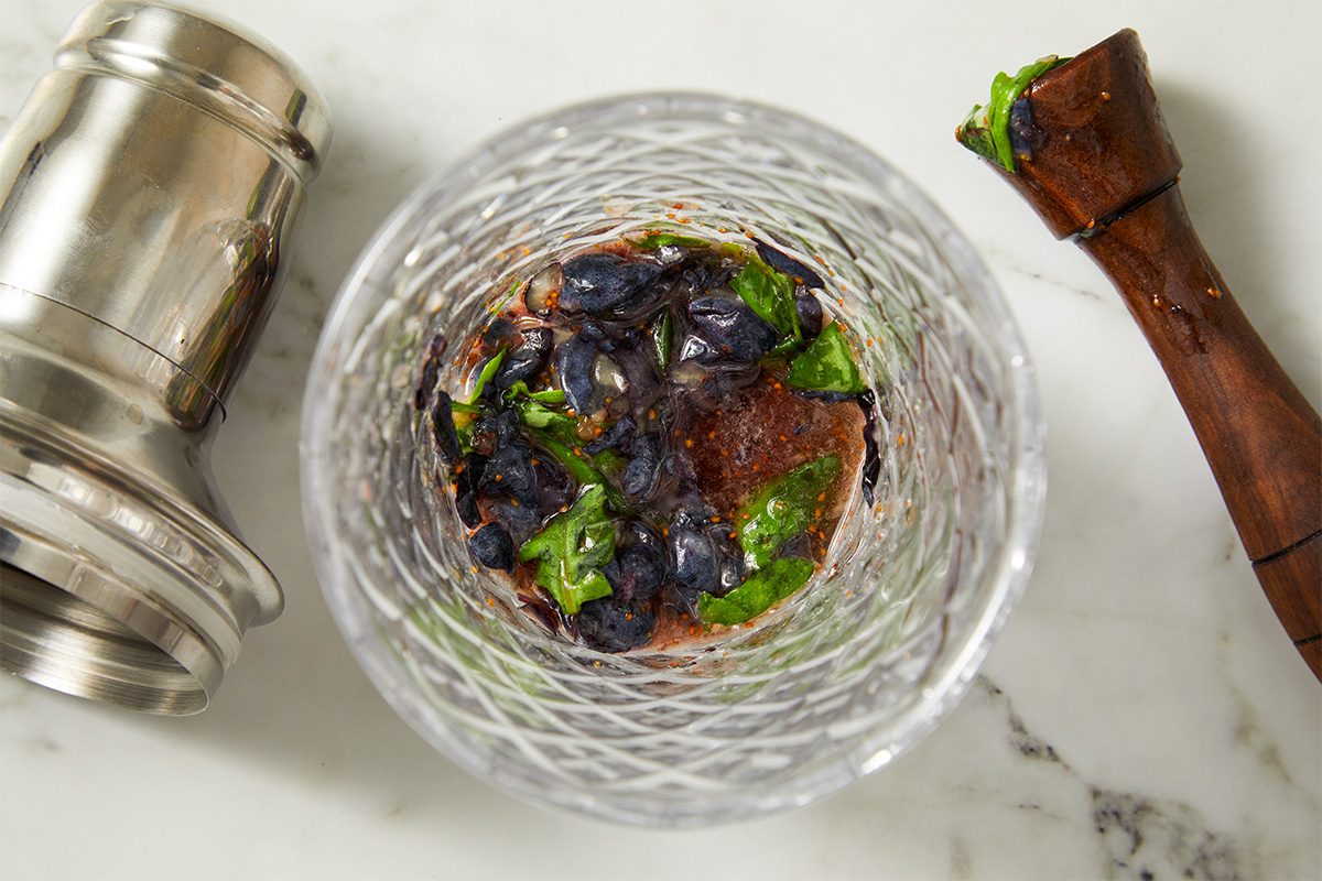 Overhead horizontal image of muddling blueberries and basil in a glass with a muddler and cocktail shaker. The dark berries and green leaves sit on top of a brown liquid.