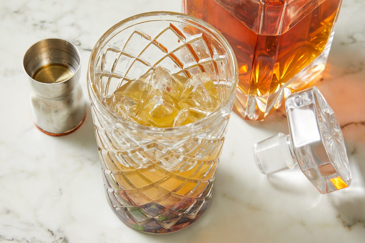 Eye Level horizontal image of a cocktail glass filled with ice and a tan-colored liquid, sitting on a white marble surface with a metal cocktail shaker on the side. A glass bottle and its stopper sit to the right of the cocktail glass.