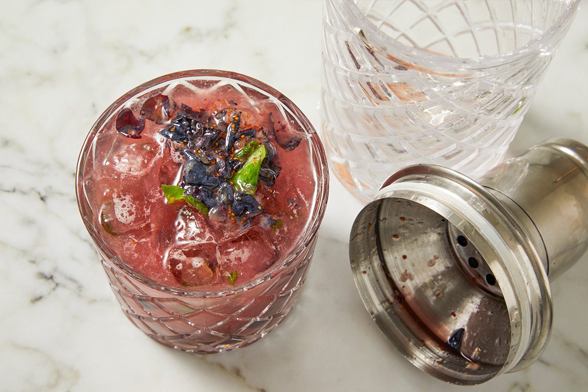 Overhead horizontal image of a cocktail in a patterned glass with ice, dark berries, and mint garnish. A second empty glass and a metal cocktail shaker are to the side on a white marble surface.