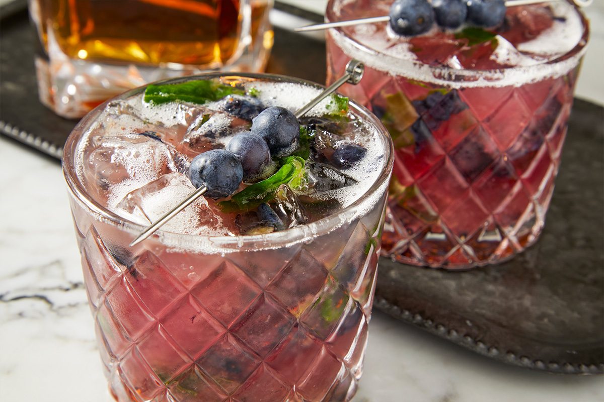 Eye Level horizontal image of a pair of Blueberry Mojitos in textured glasses, garnished with mint sprigs and blueberries on a metal cocktail skewer. One glass of whiskey is in the background, on a dark metal tray.